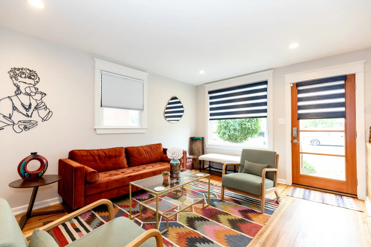 The hero image showcases a vibrant, sunlit living area featuring a striking orange sofa, a colorful patterned rug, a sleek glass coffee table, and playful, artistic wall decor. The space is framed by stylish window shades that softly filter the natural light, creating a cozy, creative atmosphere perfect for gathering with friends or relaxing with a good book.