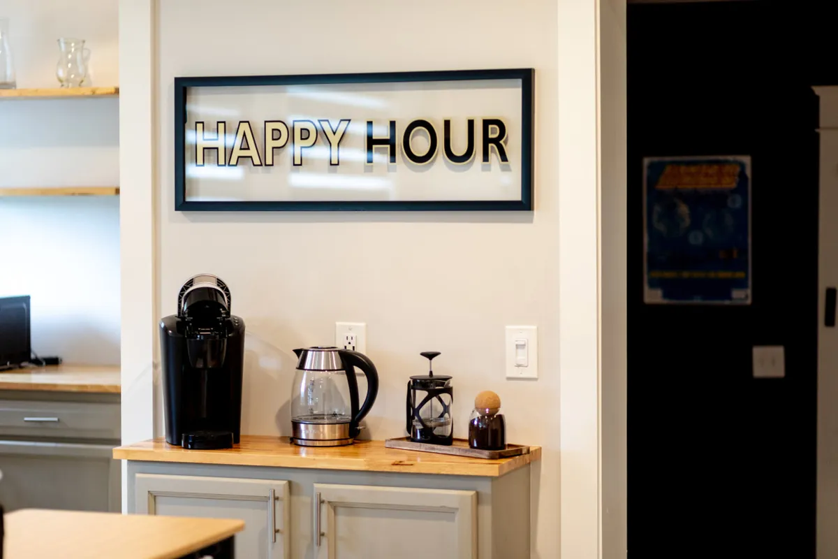 A cheerful coffee station featuring a 'HAPPY HOUR' sign, a coffee maker, coffee pot, and wooden countertop. The setup is designed for casual socialization and energizing mornings—a highlight of the curated coffee bar mentioned in the section.