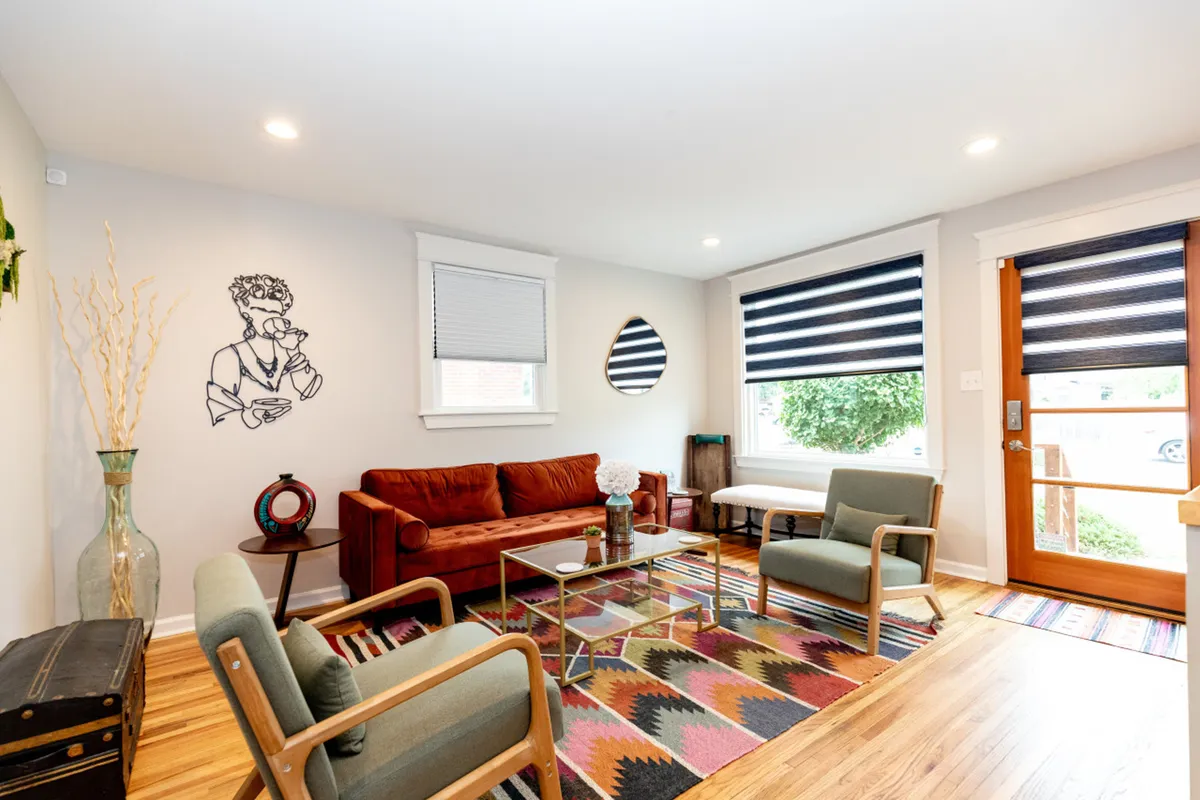 A vibrant, welcoming living area with a large artistic wall mural, colorful area rug, and mid-century modern furniture. The room is brightly lit by natural light streaming through a big window, and the space is clearly designed for group gatherings—ideal for socializing, relaxing, or playing board games.