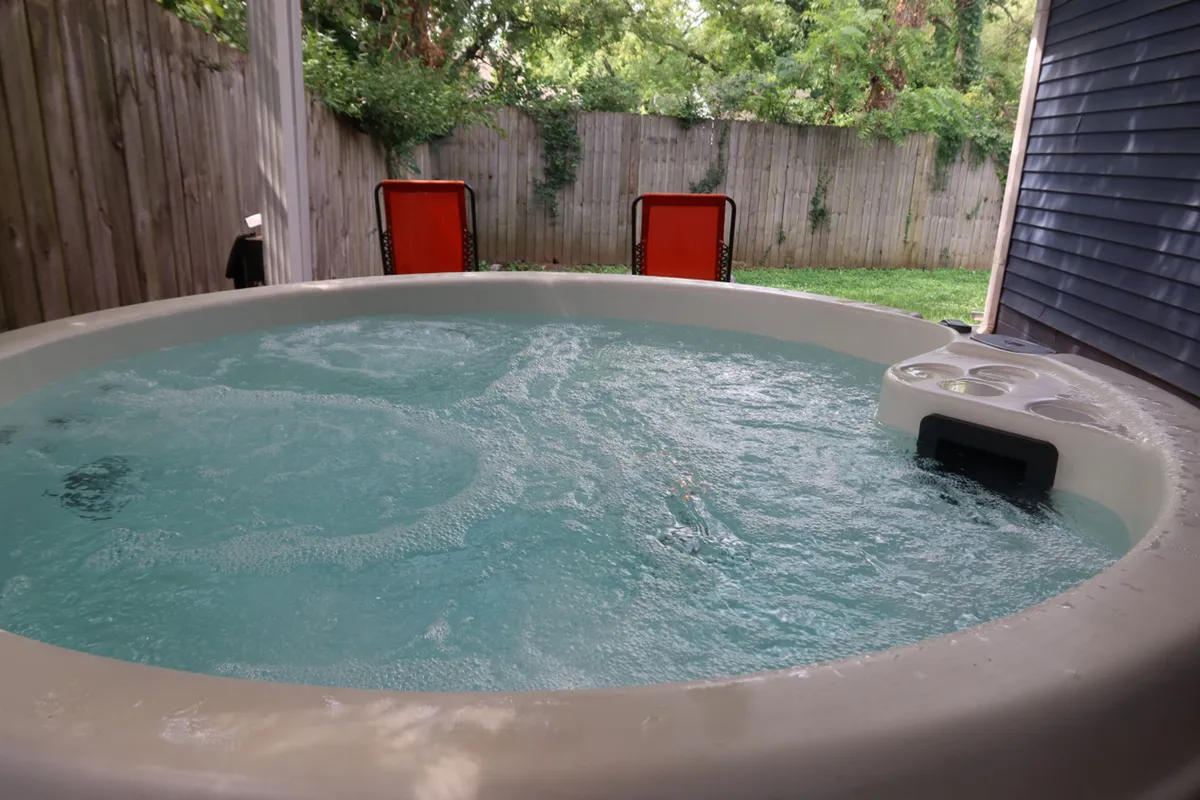 A tranquil backyard scene with a bubbling hot tub as the focal point, surrounded by green grass, orange chairs for lounging, and enclosed by a privacy fence. The mood is peaceful, inviting relaxation and socializing outdoors.