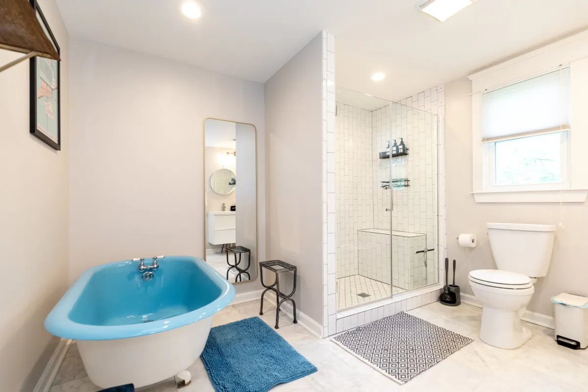 A modern bathroom with a striking blue clawfoot soaking tub, a glass-enclosed walk-in shower, a large mirror, and a stylish vanity. The room is filled with natural light, evoking a spa-like, rejuvenating mood.