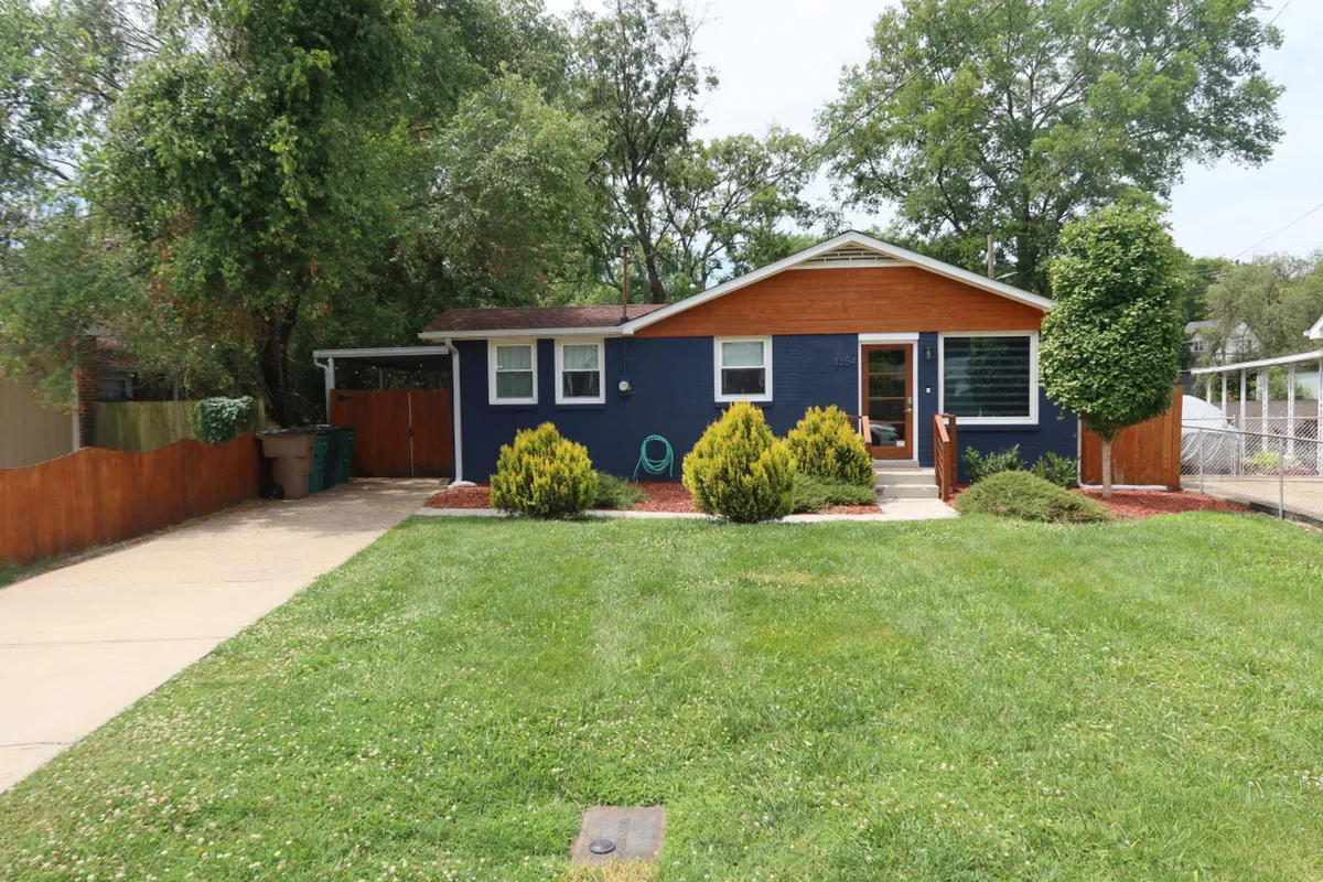 A cheerful exterior view of the home featuring a blue facade, inviting wooden accents, and a green, landscaped pathway leading directly to the front door. The image suggests ample private driveway parking and a welcoming entrance.